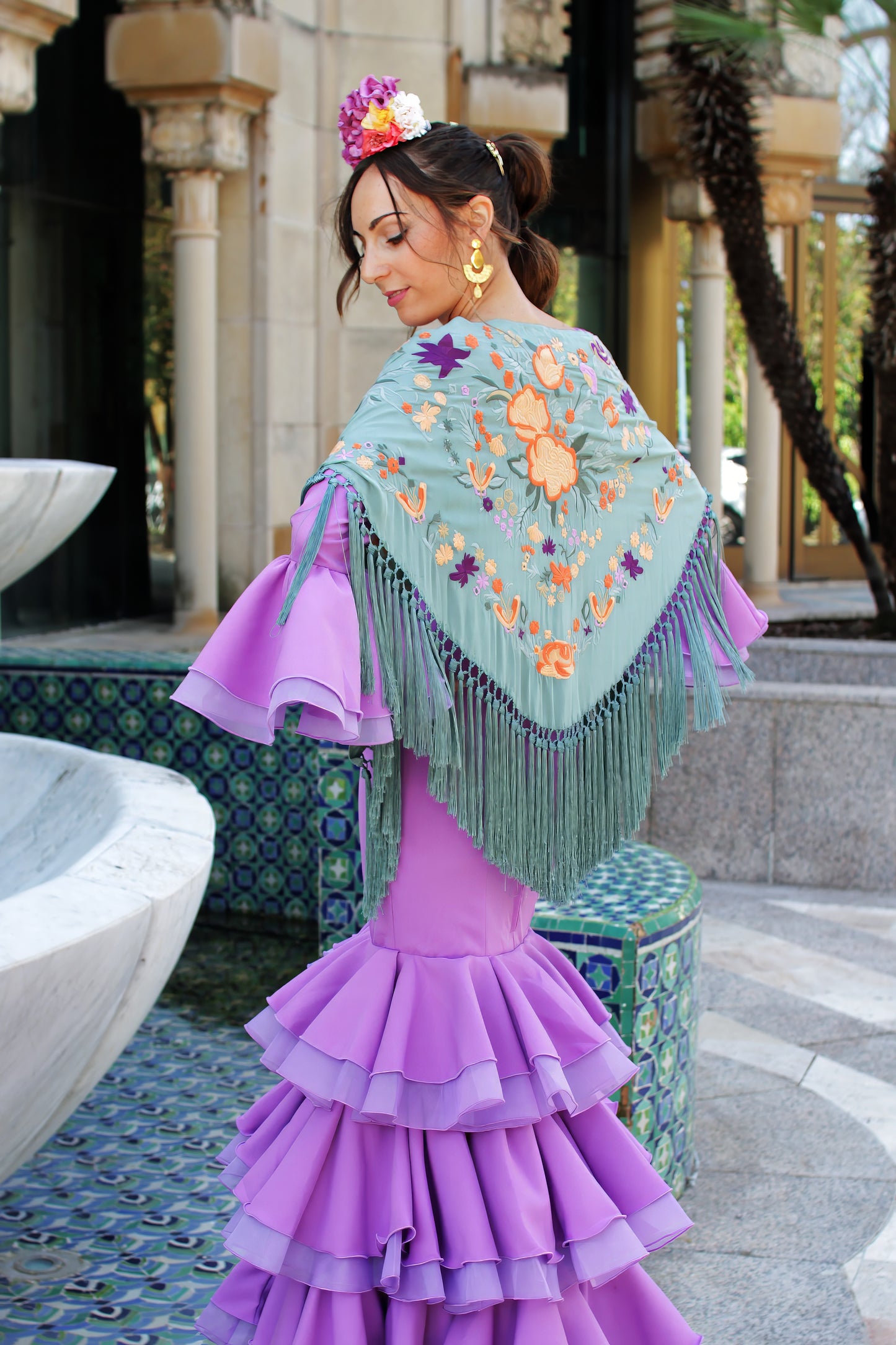 El mantón verde jabonoso Noa, con flores en naranja y morado, es perfecto para añadir frescura y originalidad a tu look flamenco.