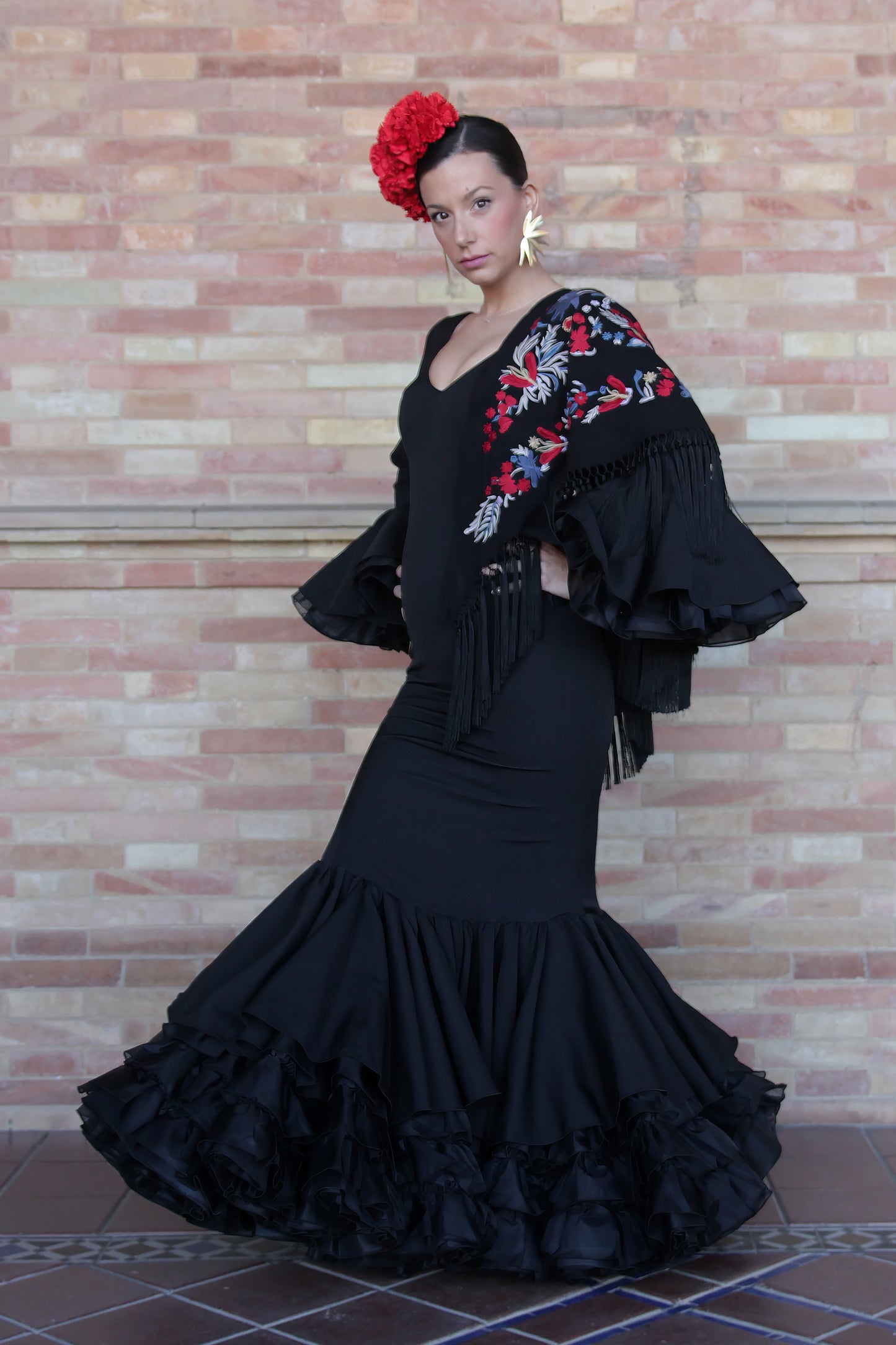 El traje de flamenca negro Clavel, complementado con mantón negro, flor roja, ramillete Rojo Pasión y pendientes dorados Hinojos, te ofrece un estilo flamenco sofisticado y vibrante para la Feria de Abril.