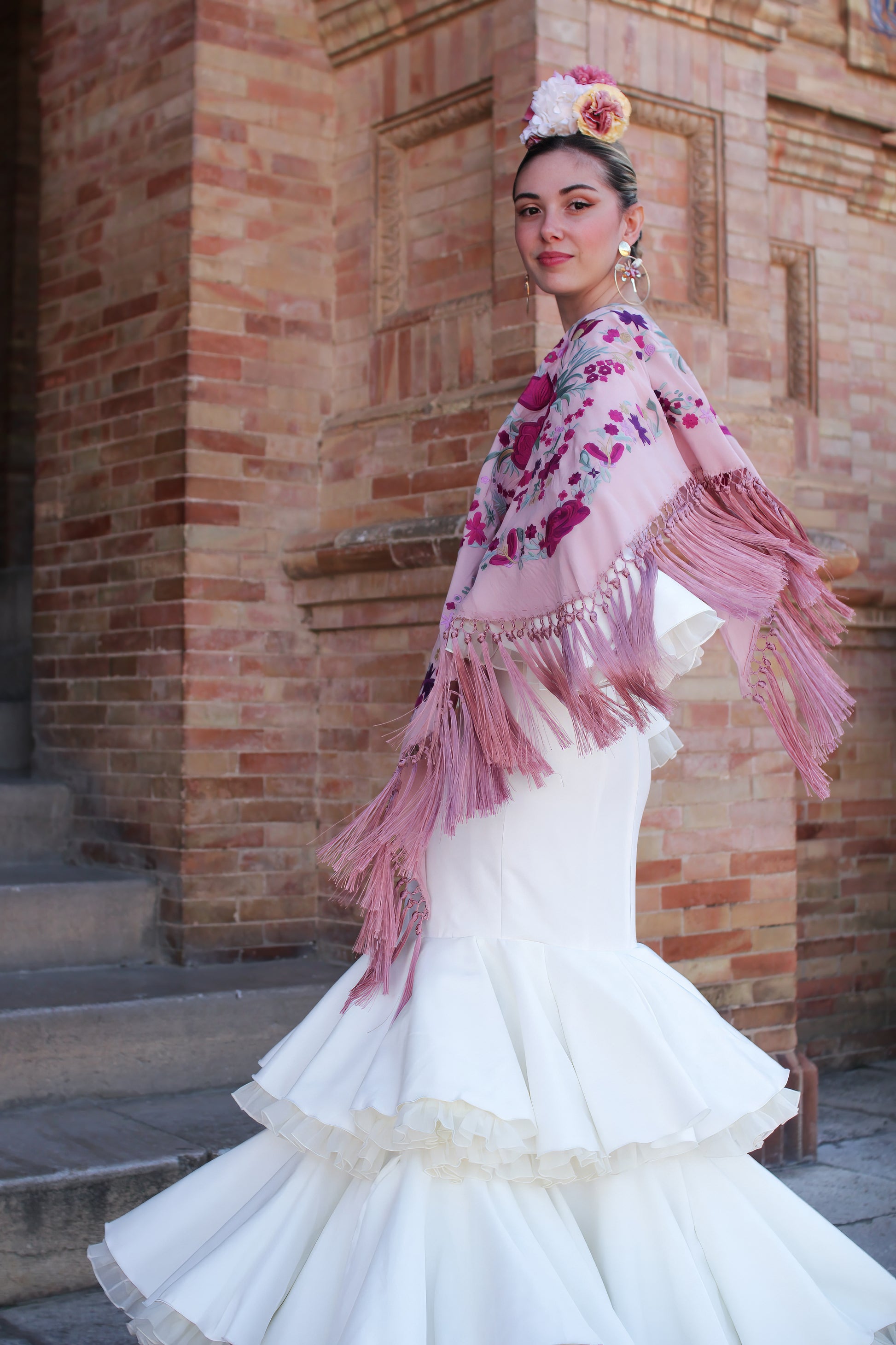 Luce la elegancia del traje de flamenca marfil Jazmin, acompañado de un manton rosa con flor buganvilla, ramillete Duende y pendientes dorados Jara, para un look flamenco sofisticado y lleno de personalidad.