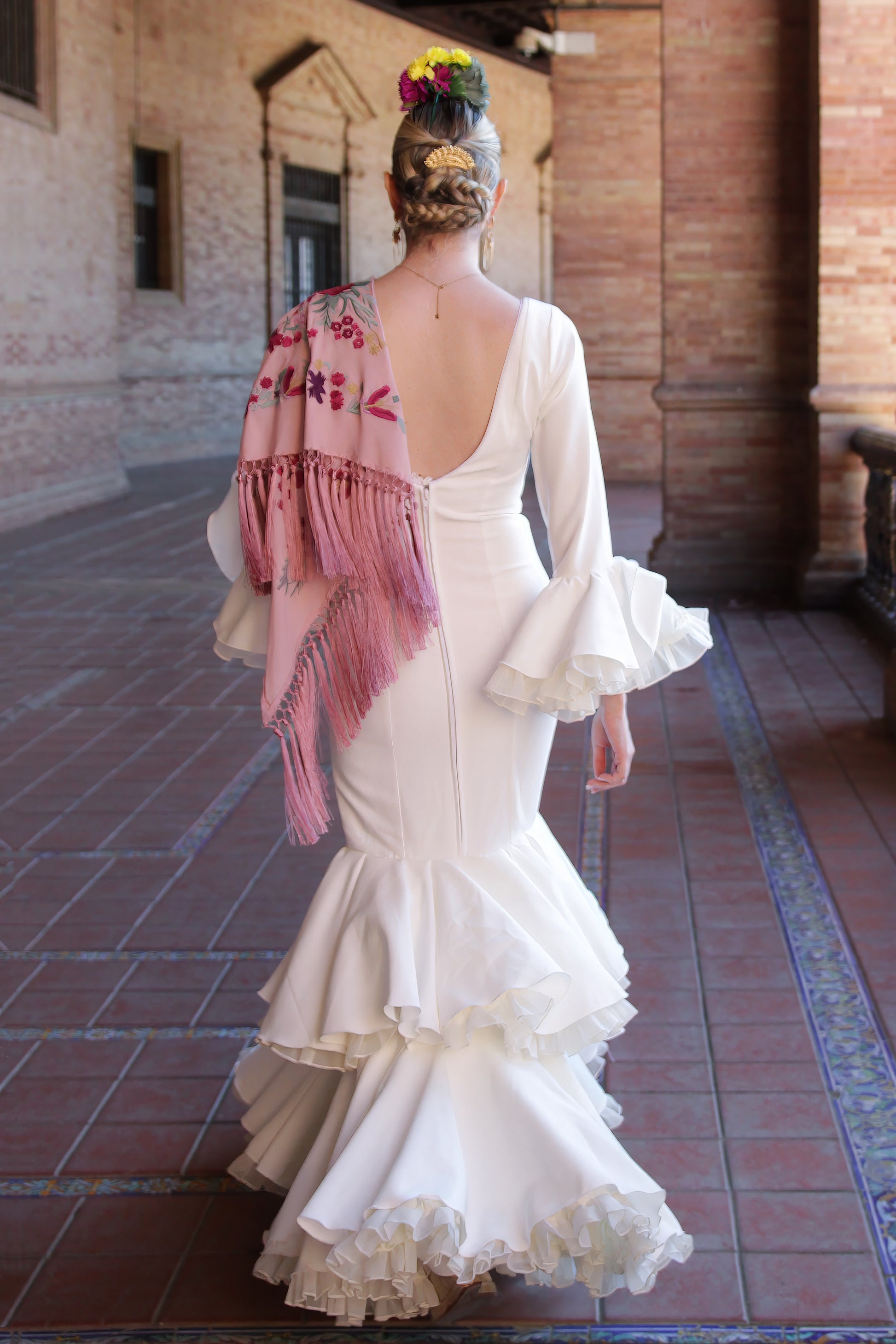 El traje de flamenca marfil Jazmin es perfecto para la Feria de Abril, con volantes delicados y un manton rosa con flor buganvilla. Complementado con un ramillete Duende y pendientes dorados Jara para un look completo y vibrante.