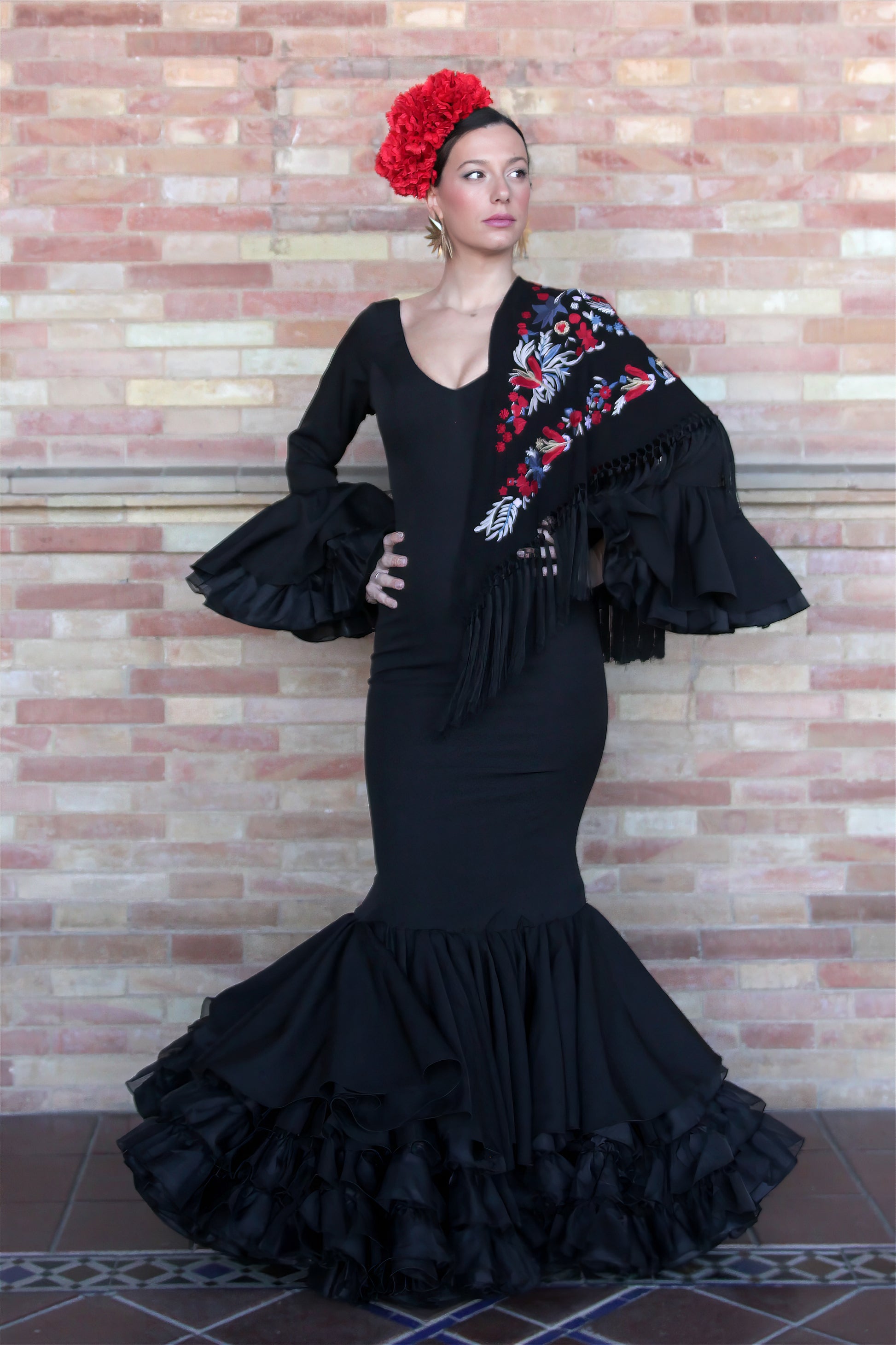 Destaca en la Feria de Abril con el vestido de flamenca negro Clavel, acompañado de un mantón negro con flor roja, ramillete Rojo Pasión y pendientes dorados Hinojos. Un conjunto lleno de elegancia y movimiento.