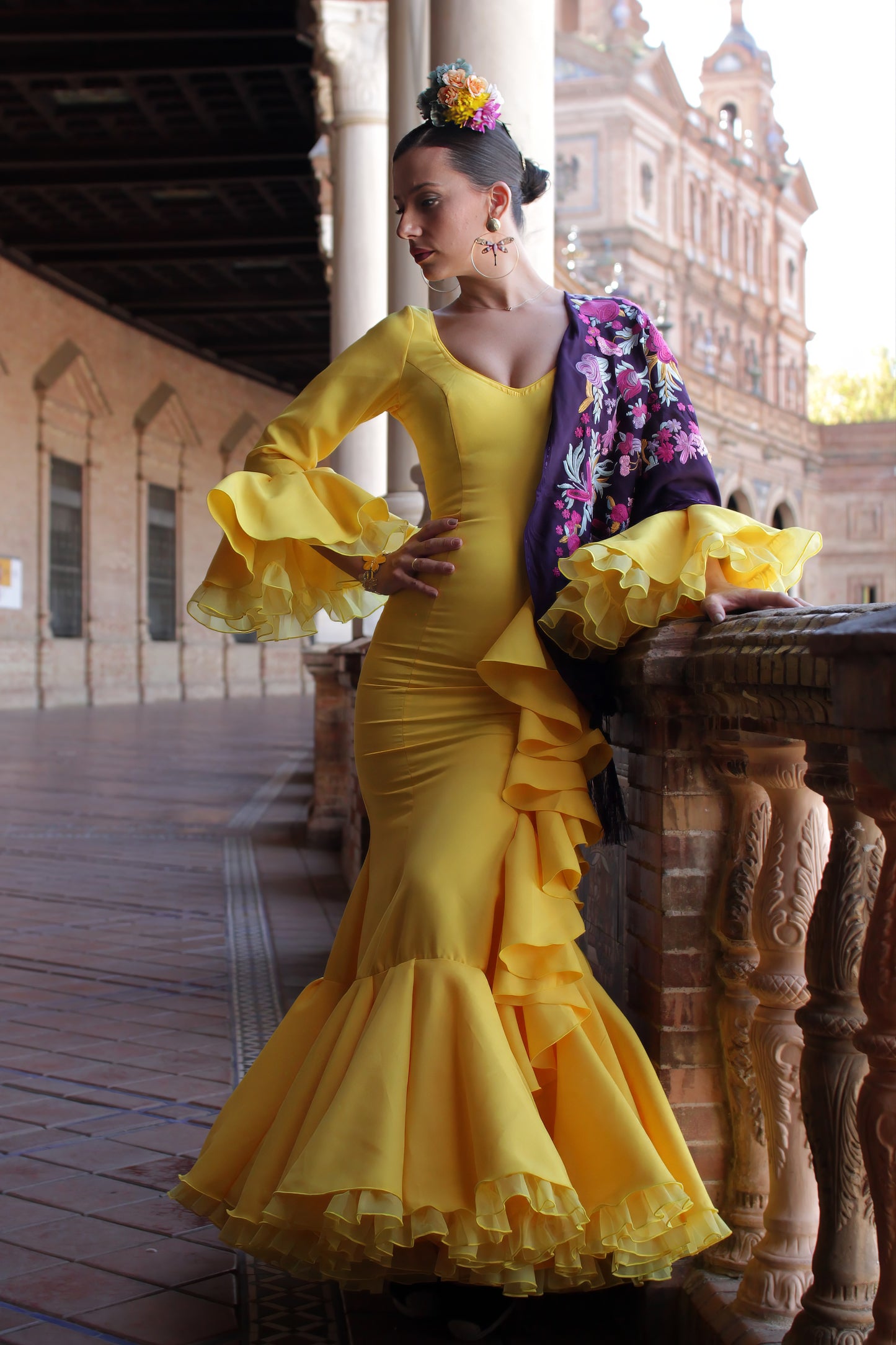 Destaca en la Feria de Abril con el traje de flamenca amarillo Azahar, acompañado de un mantón morado con patrón buganvilla y amarillo, ramillete Brisa y pendientes dorados Jara. Look lleno de alegría y elegancia.