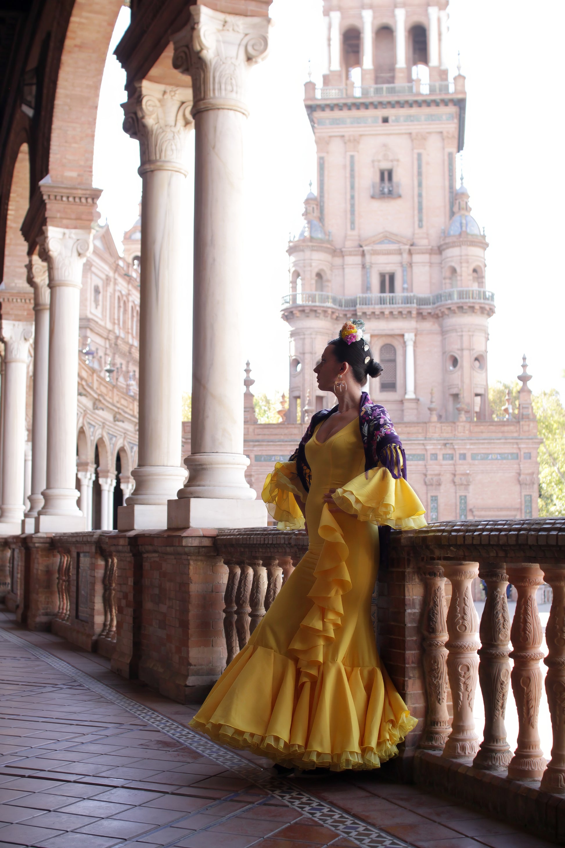 Completa tu look para la Feria con el vestido amarillo Azahar, mantón morado con detalles en buganvilla, ramillete Brisa y pendientes dorados Jara. Mostrando la combinación perfecta para la feria.
